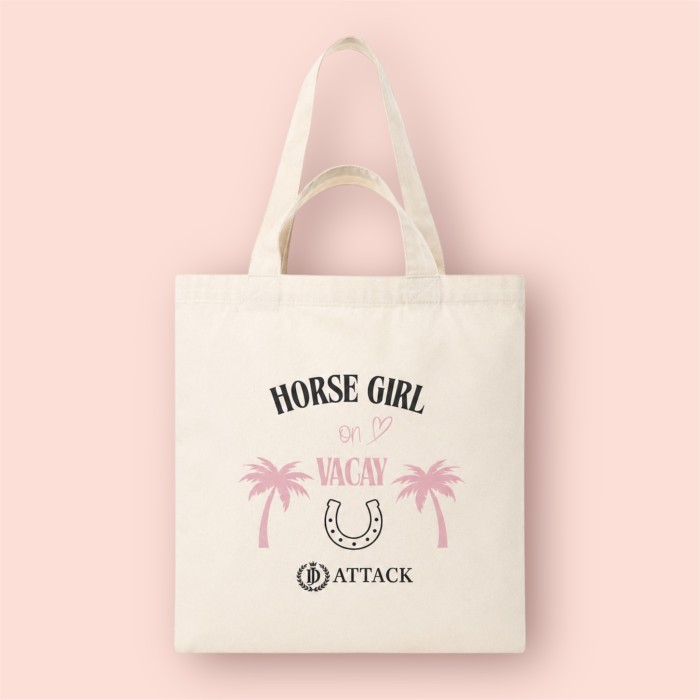 Ekologiczna torba bawełniana Vacay od JD Attack w kolorze ecru na jasnoróżowym tle. Widoczny czarno-różowy nadruk Horse Girl on Vacay z palmami, podkową i logo marki oraz praktyczne, podwójne uchwyty na ramię i do ręki.