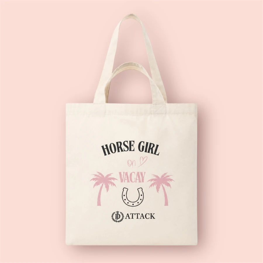 Ekologiczna torba bawełniana Vacay od JD Attack w kolorze ecru na jasnoróżowym tle. Widoczny czarno-różowy nadruk Horse Girl on Vacay z palmami, podkową i logo marki oraz praktyczne, podwójne uchwyty na ramię i do ręki.