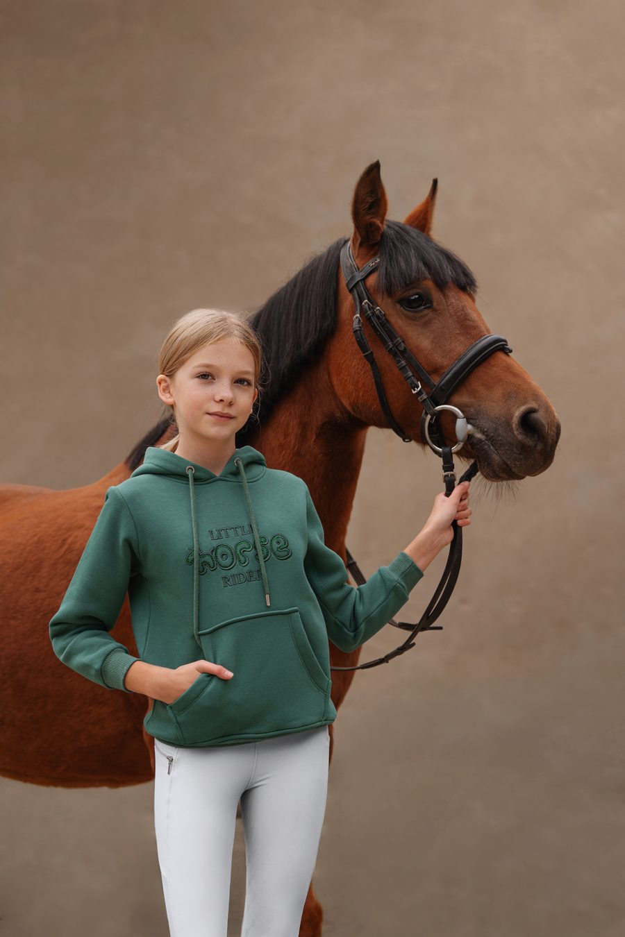 Dziewczynka w bluzie z kapturem z napisem Little Horse Rider i jasnych legginsach jeździeckich obok konia